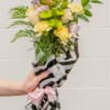 Hand-tied bouquet of pink lilies, yellow carnations, and green mums in striped wrap with a pink bow