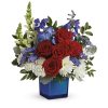 Patriotic arrangement of red roses, red carnations, white mums and blue flowers in a blue glass cube vase