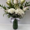 Tall arrangement of white roses, lilies, and carnations in a green glass vase with a white bow
