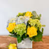 Compact arrangement of yellow roses and blue and green hydrangeas in a rustic wooden cube vase