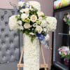 White floral cross arrangement with cream roses and blue accents on an easel