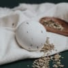 White bath bomb with herb flecks on a folded towel beside a wooden spoon of dried leaves.