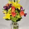 Bouquet of yellow lilies, orange roses, and purple flowers in a clear glass vase