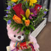Mixed bouquet in a glass vase with a pink teddy bear and ribboned gift.