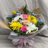 Hand-tied bouquet of pink roses, yellow daisies, and white mums wrapped in kraft paper with a pink bow.