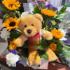 Stuffed teddy bear nestled among yellow sunflowers, white carnations, and pink and purple daisies.