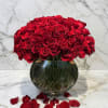Round arrangement of red roses in a clear glass bowl vase with scattered rose petals.