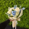 Hand-tied bouquet of white roses and blue flowers in cream wrap with ribbon, held against a green wall.
