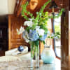 Tall floral arrangement with blue hydrangeas, white orchids, and green hanging garlands in a glass vase