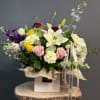 Colorful mixed flower arrangement with lilies, roses, and carnations in a white box on a wood stand.