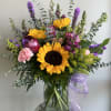 Tall mixed arrangement with sunflowers, pink roses, and purple flowers in a clear glass vase