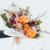 Hand-tied bouquet of orange and yellow blooms with burgundy accents wrapped in white paper.