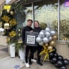 Two people stand beside a balloon cluster and holiday storefront decor.