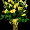 Tall yellow and green floral arrangement with lilies, roses, and daisies in a patterned cylinder vase with bow