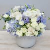 Low white ceramic vase with white roses, blue hydrangeas, and light blue flowers in a rounded arrangement