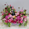 Low pink floral arrangement with roses and tulips surrounding a bottle of champagne in a white container.