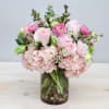 Round arrangement of pink hydrangeas, roses, and ranunculus in a clear glass vase with pebbles.