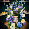 Tall floral arrangement with white roses, blue delphinium, and purple mums around a candle
