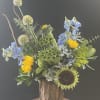 Mixed bouquet with sunflowers, blue flowers, and green textured blooms in a rustic wood vase