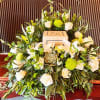 Cremation urn surrounded by white roses, lilies, green mums, and a succulent on a wooden casket