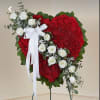 Heart-shaped red carnation standing spray with white roses, white ribbon bow, and greenery on an easel.