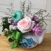 Pastel pink and purple flower arrangement in a painted ceramic teapot with green bow and tilted lid