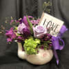 Purple and lavender floral arrangement in a white ceramic vase with a ribbon and "LOVE" sign