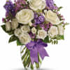 White roses and purple flowers arranged in a glass vase with a purple ribbon.
