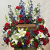 Large funeral spray with red roses, white lilies, and blue delphinium around a wooden urn