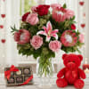 Pink and red bouquet with lilies and protea in a glass vase beside chocolates and a red teddy bear