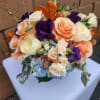 Compact arrangement of peach and cream roses, purple lisianthus, and blue hydrangea with orange leaves