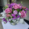 Compact arrangement of pink roses and purple flowers in a round glass vase