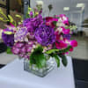 Compact arrangement of purple and pink flowers in a clear glass cube vase.