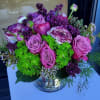 Compact arrangement of pink roses, purple blooms, and green hydrangeas in a clear glass vase