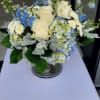 Low round arrangement of cream roses, blue delphinium, hydrangeas, and white mums in a clear glass vase