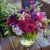 Low glass vase of fuchsia roses, purple blooms, and mixed pink flowers with a card tucked at the back.
