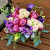 Low arrangement of pink and cream roses with purple flowers in a clear vase on a wooden bench