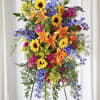 Tall standing spray with sunflowers, orange lilies, blue delphinium, and bright pink blooms on an easel