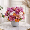 Low round arrangement of pink roses, orchids, and orange spray roses in a white pot on a marble table
