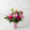 Pink roses, lilies, and daisy-style blooms in a pink patterned vase with a heart ribbon