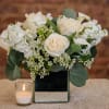 White rose arrangement in a mirrored square vase with a small candle beside it