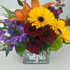 Bouquet of yellow gerbera daisies, red roses, orange lilies, and purple flowers in a glass vase