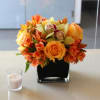 Orange and yellow floral arrangement in a black square vase beside a candle