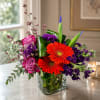 Bright mixed bouquet with orange gerbera daisies, pink roses, and purple flowers in a glass vase