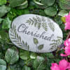 Decorative stone engraved with the word Cherished among green leaves and pink flowers