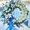Standing white and blue funeral wreath with lilies and ribbon on a metal easel.