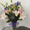 Mixed bouquet of lilies, irises, and pink flowers in a glass vase with a purple ribbon