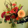 Hand-tied bouquet of orange gerbera, red mums, carnations, and roses with tall filler stems, held against a pale wall