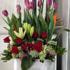 Tall floral arrangement with red roses, purple tulips, and white lilies in a white container
