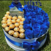 Blue roses and gold-wrapped chocolates arranged in a round gift box with a ribbon.